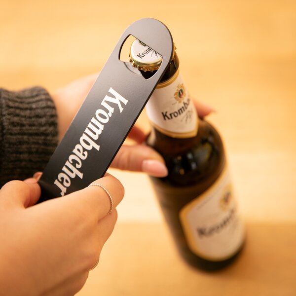 Hand öffnet Krombacher Bierflasche mit schwarzem Flaschenöffner mit weißem Krombacher Schriftzug.