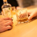 Zwei Hände halten Kristallgläser mit Bierbrand auf einem Holztisch.