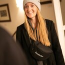 Frau trägt schwarze Krombacher Bauchtasche über schwarzem Mantel und grauem Pullover mit weißer Strickmütze.