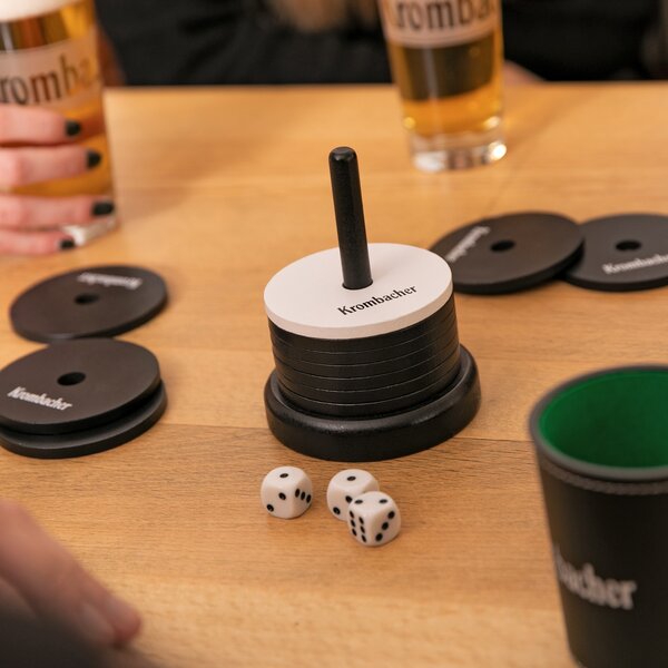 Krombacher Schockbesteck Spielscheiben mit schwarzer und weißer Farbe und Krombacher Schriftzug, Würfel und zwei Biergläser auf Holztisch.