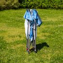 Zusammengeklappter blauer und weißer Pavillon auf einer grünen Wiese mit Büschen im Hintergrund.
