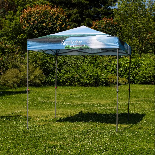 Blauer Pavillon mit Krombacher-Logo und Schriftzug 'Eine Perle der Natur' auf einer grünen Wiese vor Bäumen.