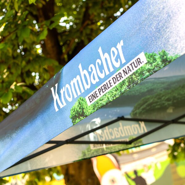 Blauer Pavillon mit weißem Schriftzug 'Krombacher' und Slogan 'Eine Perle der Natur' unter Baumkronen.