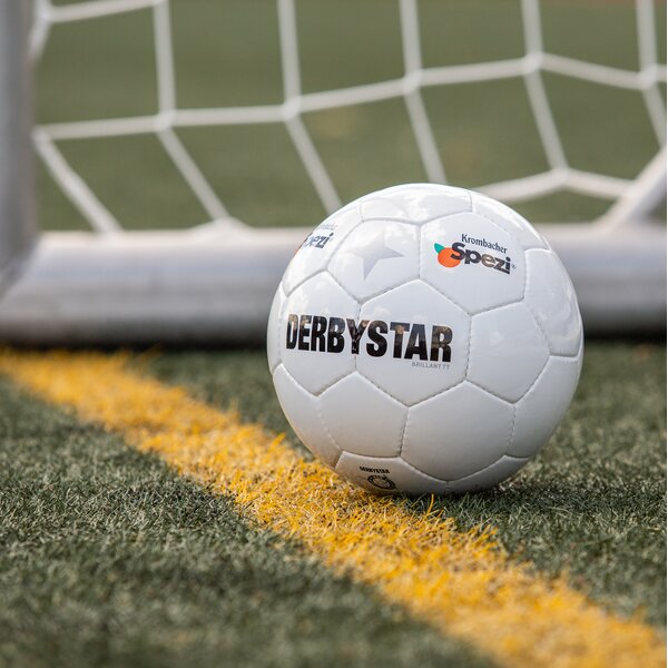 Weißer Fußball mit schwarzem Schriftzug 'DERBYSTAR' und Logos von Krombacher Spezi, liegt hinter der Torlinie auf grünem Rasen.