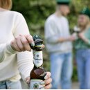 Hand öffnet Krombacher Pils Bierflasche mit Schlüsselanhänger in Form einer grünen Schützenkappe.