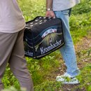 Zwei Personen tragen eine Krombacher Kühltasche mit Bierkasten auf einem Waldweg.