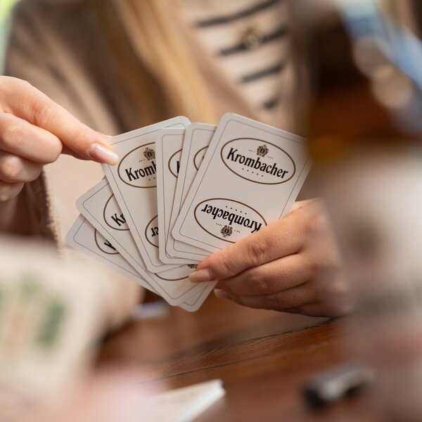 Person hält fünf Skat Spielkarten mit dem Krombacher-Logo in der Hand.