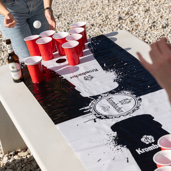 Person spielt Beerpong mit roten Bechern auf Krombacher-Matte auf weißem Tisch im Freien, Krombacher Pils Flasche daneben.