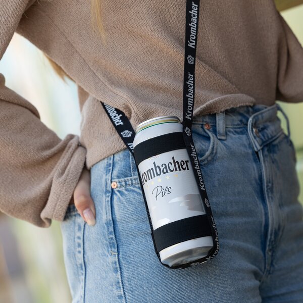 Krombacher Pils Dose in schwarzem Getränkehalter mit Krombacher-Schriftzug, getragen an schwarzem Band um die Schulter einer Person in beigem Pullover und blauer Jeans.
