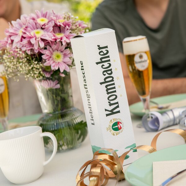 Krombacher Bierverpackung mit Schrift 'Lieblingsflasche' auf gedecktem Tisch mit Blumen, Bierglas und Personen im Hintergrund.