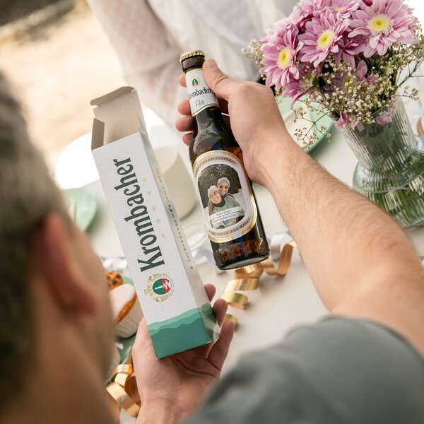 Person hält eine Krombacher Pils Flasche mit individuellem Etikett und passende 1er Verpackung, daneben ein Blumenstrauß mit rosa Chrysanthemen.