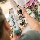 Person hält eine Krombacher Pils Flasche mit individuellem Etikett und passende 1er Verpackung, daneben ein Blumenstrauß mit rosa Chrysanthemen.