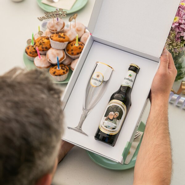 Person hält geöffnete Geschenkbox mit Krombacher Pils Flasche mit individuellem Etikett und passendem Glas mit goldfarbenem Rand.