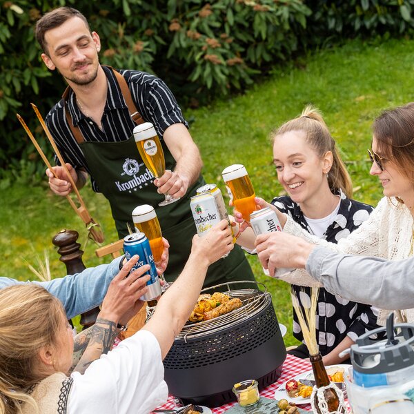 Grillmeister in Krombacher-Grillschürze mit Bierglas, Freunde stoßen mit Krombacher an, Grill mit Fleisch und Gemüse.