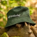 Dunkelgrüner Bucket Hat mit Krombacher Logo, liegend auf einem Baumstamm.