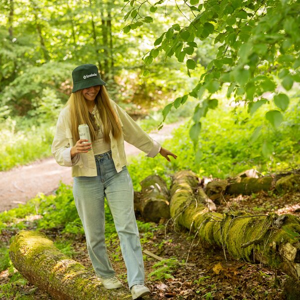 Frau mit langem Haar trägt den Krombacher Bucket Hat, balanciert auf einem moosbedeckten Baumstamm im Wald.