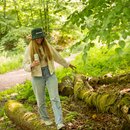 Frau mit langem Haar trägt den Krombacher Bucket Hat, balanciert auf einem moosbedeckten Baumstamm im Wald.