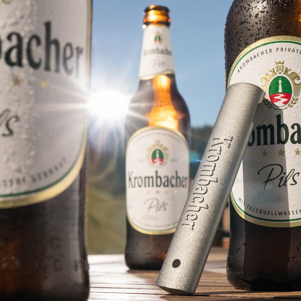 Flaschenöffner neben Krombacher Bierflaschen auf einem Tisch im Sonnenlicht.