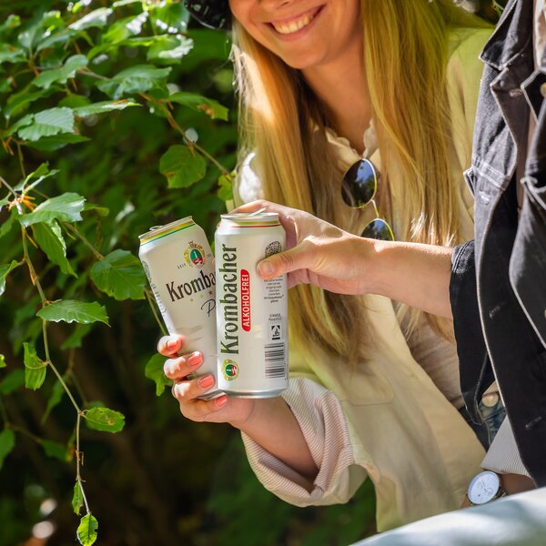 529_krombachershop_0,5_AF_Mood_004 Zwei Personen stoßen mit Dosen Krombacher Pils alkoholfrei 24x0,5L Dose an im Freien vor grünem Laub.