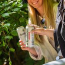 529_krombachershop_0,5_AF_Mood_004 Zwei Personen stoßen mit Dosen Krombacher Pils alkoholfrei 24x0,5L Dose an im Freien vor grünem Laub.