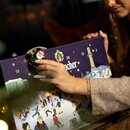 7483_krombachershop_Adventskalender_Mood_002 Hand einer Frau mit beigem Pullover und goldfarbenem Armband holt eine kleine Weihnachtsmannfigur aus einem Adventskalender mit winterlicher Illustration und Zahlen.