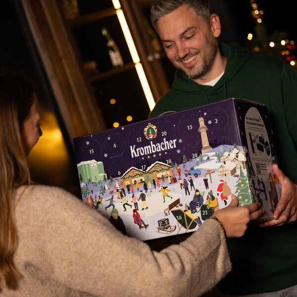 Personen mit Krombacher Adventskalender. Zwei Personen halten gemeinsam einen Krombacher Adventskalender mit winterlicher Illustration und nummerierten Türchen.