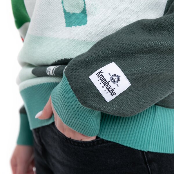 Nahaufnahme eines dunkelgrünen Ärmels mit einem weißen Krombacher-Logoetikett an einem Pullover, Hand in Hosentasche.