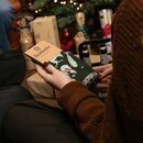 Person hält grüne Weihnachtssocken mit Krombacher-Logo und Weihnachtsmotiven vor einem geschmückten Weihnachtsbaum und Geschenken.