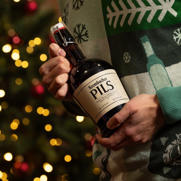 Person hält eine Flasche Krombacher Pils mit Bügelverschluss vor einem Weihnachtsbaum mit Lichterketten, trägt grünen Pullover mit Schneeflockenmuster.