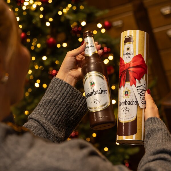 Person hält eine braune Krombacher Pils Flasche aus Schokolade und eine Geschenkverpackung mit roter Schleife und Aufschrift 'Schokoladen Flasche'.