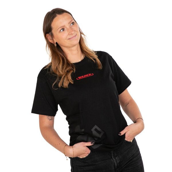 Frau trägt schwarzes T-Shirt mit rotem Schriftzug 'Rolinch' und dunkle Jeans, Hände in den Hosentaschen.