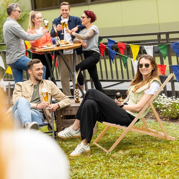 Gruppe von Menschen sitzt und steht im Garten, zwei Personen auf Liegestühlen mit Getränken, andere an einem Stehtisch mit Bier.
