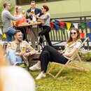 Gruppe von Menschen sitzt und steht im Garten, zwei Personen auf Liegestühlen mit Getränken, andere an einem Stehtisch mit Bier.