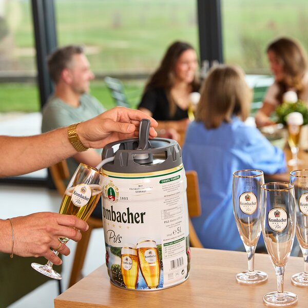 Hand gießt Krombacher Pils aus einem Frischefässchen in ein Glas, im Hintergrund sitzen mehrere Personen an einem Tisch