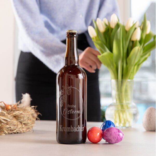 Braune Bierflasche mit eingraviertem Schriftzug 'Frohe Ostern' und Krombacher Logo auf einem Tisch neben bunten Ostereiern und einem Glas mit Tulpen, im Hintergrund eine Person in blauem Pullover