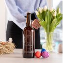 Braune Bierflasche mit eingraviertem Schriftzug 'Frohe Ostern' und Krombacher Logo auf einem Tisch neben bunten Ostereiern und einem Glas mit Tulpen, im Hintergrund eine Person in blauem Pullover