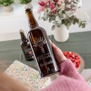 Braune Krombacher Flasche mit Gravur 'Für die beste Mama' und Gerstenstraußmotiv, gehalten von einer Person in rosa Pullover vor unscharfem Hintergrund mit Blumen und Teller mit roten Früchten