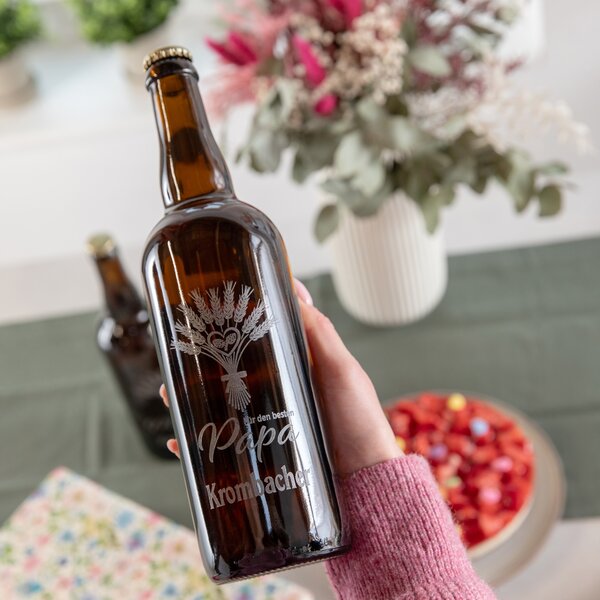 Hand hält eine braune Bierflasche 0,75 L mit Gravur 'Für den besten Papa' und Krombacher Logo vor unscharfem Hintergrund mit Blumen und Schale voller Süßigkeiten