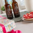 Zwei braune Krombacher-Flaschen mit Gravur "Für den besten Papa" und "Für die beste Mama" auf einem Tischläufer neben einem Erdbeerkuchen und einem Geschenk mit pinkem Band.