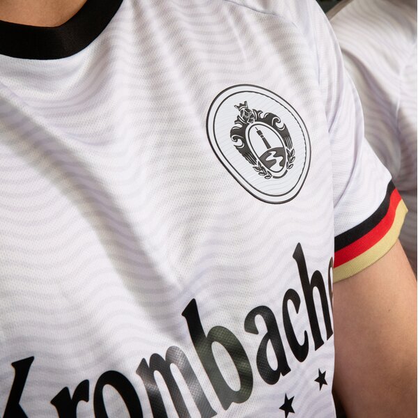 Ausschnitt eines weißen Trikots mit schwarzem Kragen, einem Wappen auf der Brust und dem Schriftzug 'Krombacher' sowie einem Ärmel mit schwarz-rot-goldenem Rand