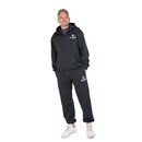 Mann steht mit Händen in den Taschen und trägt schwarzen Kapuzenanzug mit weißem Krombacher-Logo auf Brust und Hose sowie weiße Sportschuhe