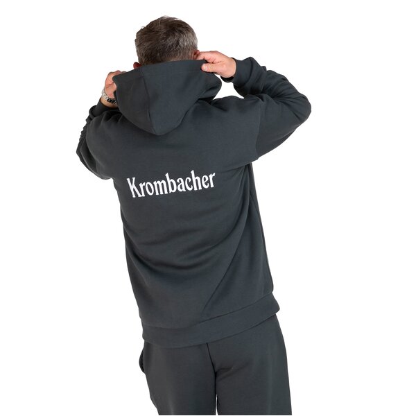 Rückansicht eines Mannes, der eine Kapuzenjacke mit dem weißen Schriftzug 'Krombacher' trägt und die Kapuze mit beiden Händen hält