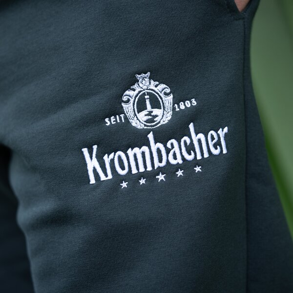 Nahaufnahme eines dunkelgrünen Kleidungsstücks mit weißer Stickerei des Schriftzugs 'Krombacher' und einem Wappen mit der Aufschrift 'SEIT 1803'