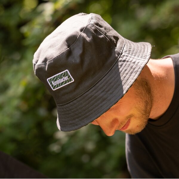 Mann mit dunkelgrünen Bucket Hat mit aufgenähtem Krombacher-Logo blickt nach unten