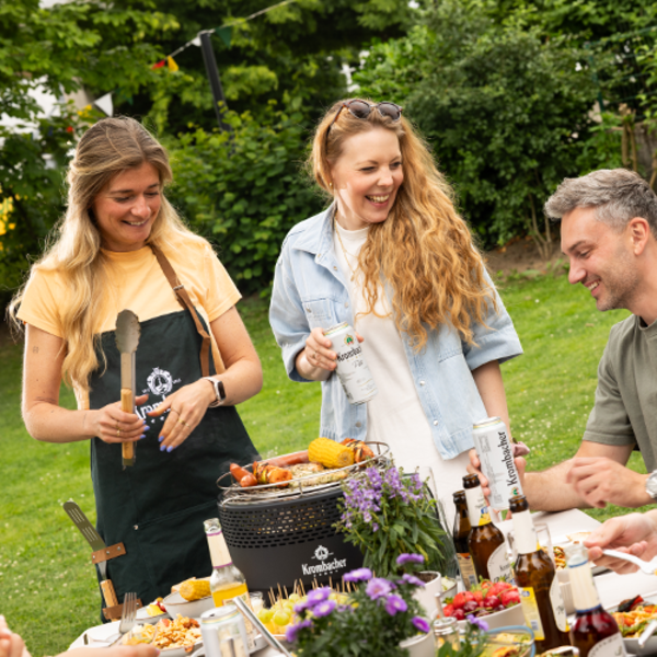 Drei Personen stehen lachend um einen kleinen Grill im Garten, eine Frau hält Grillzange, eine andere eine Getränkedose mit der Aufschrift 'Krombacher'