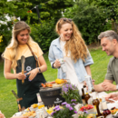 Drei Personen stehen lachend um einen kleinen Grill im Garten, eine Frau hält Grillzange, eine andere eine Getränkedose mit der Aufschrift 'Krombacher'