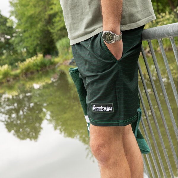Mann lehnt mit einer Hand in der Tasche an einem Geländer und trägt grüne Krombacher-Badeshorts mit Logo am Bein