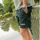 Mann lehnt mit einer Hand in der Tasche an einem Geländer und trägt grüne Krombacher-Badeshorts mit Logo am Bein