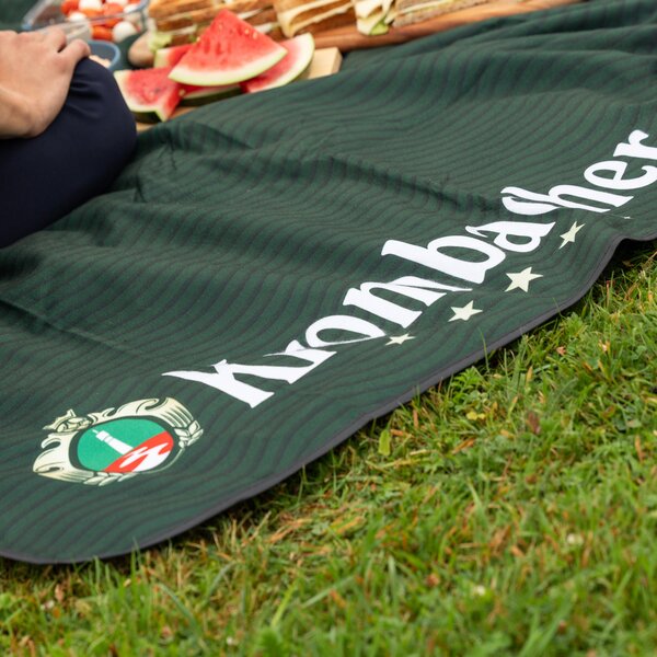 Picknickdecke mit Krombacher-Logo und Schriftzug auf Gras liegend, darauf Wassermelonenstücke und eine sitzende Person.