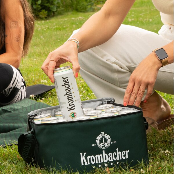 Hand nimmt eine Krombacher-Dose aus einer grünen Kühltasche mit Krombacher-Logo auf einer Wiese